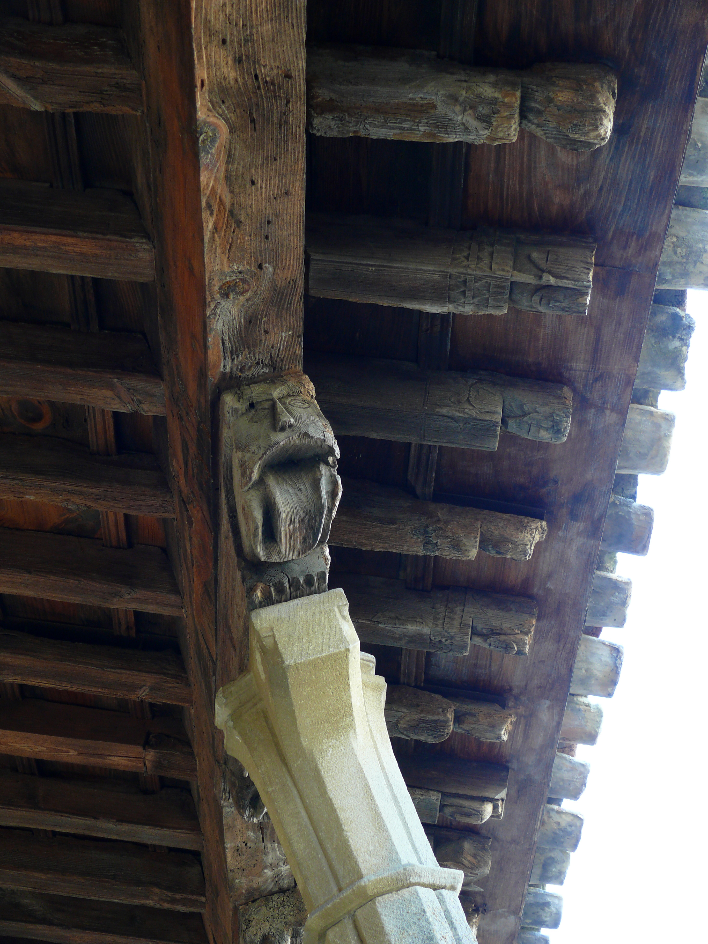 Detalle de la cubierta de la galería, toda ella de madera. Tanto las zapatas sobre los pilares, como los extremos de los cabrios en el alero, están adornados con diversas cabezas y figuras talladas.